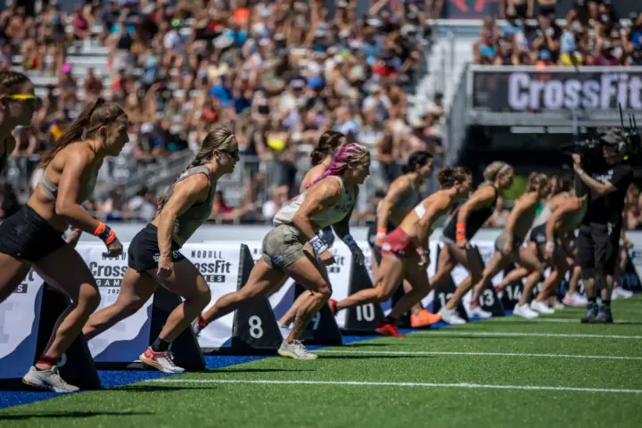 Female athletes sprinting from the start line during a CrossFit Games competition in a packed stadium.