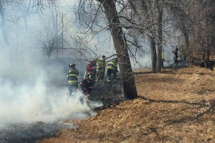 Firefighters extinguishing smoldering grass and brush under trees in a greenbelt near Pinnacle Charter High School in Thornton after a fast-spreading wildfire.