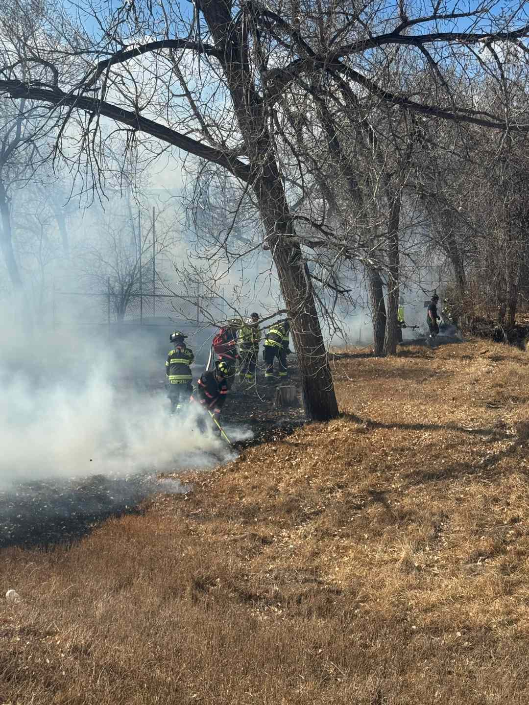 Firefighters extinguishing smoldering grass and brush under trees in a greenbelt near Pinnacle Charter High School in Thornton after a fast-spreading wildfire