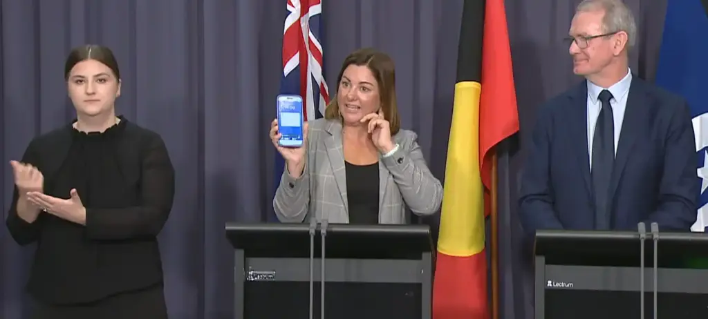 Kristy McBain holding a smartphone showing an emergency alert during an AusAlert press conference in Canberra, flanked by officials and a sign language interpreter.