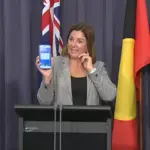 Kristy McBain holding a smartphone showing an emergency alert during an AusAlert press conference in Canberra, flanked by officials and a sign language interpreter.