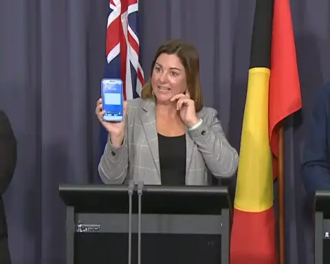 Kristy McBain holding a smartphone showing an emergency alert during an AusAlert press conference in Canberra, flanked by officials and a sign language interpreter.