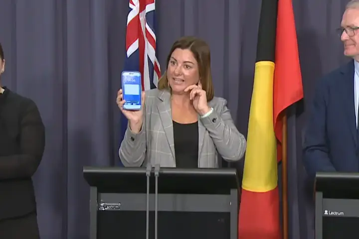 Kristy McBain holding a smartphone showing an emergency alert during an AusAlert press conference in Canberra, flanked by officials and a sign language interpreter.