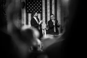 President Donald Trump seated at the podium in the U.S. Capitol chamber with two lawmakers standing behind him applauding, American flag displayed in the background during a joint session of Congress.