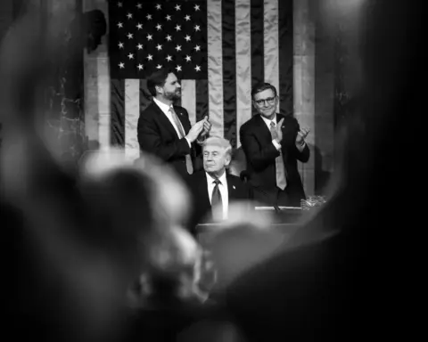President Donald Trump seated at the podium in the U.S. Capitol chamber with two lawmakers standing behind him applauding, American flag displayed in the background during a joint session of Congress.