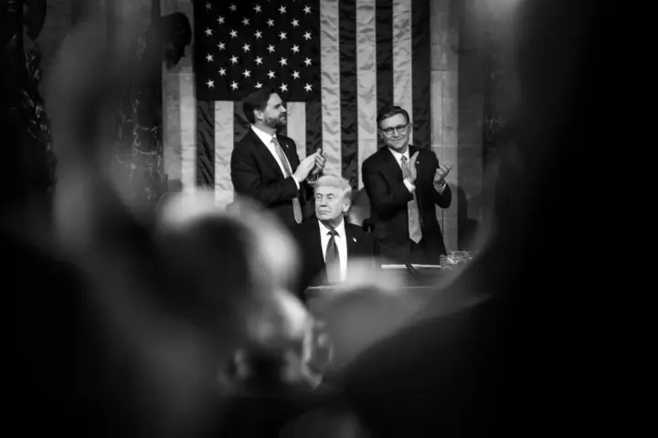 President Donald Trump seated at the podium in the U.S. Capitol chamber with two lawmakers standing behind him applauding, American flag displayed in the background during a joint session of Congress.