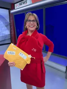 Meteorologist Jeriann Ritter in a red outfit holding a mailed package and wearing a personalized scarf in a TV studio.