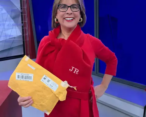 Meteorologist Jeriann Ritter in a red outfit holding a mailed package and wearing a personalized scarf in a TV studio.
