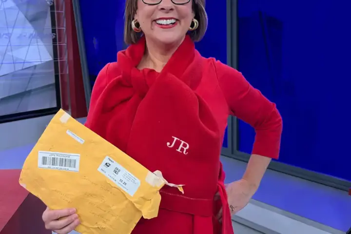 Meteorologist Jeriann Ritter in a red outfit holding a mailed package and wearing a personalized scarf in a TV studio.