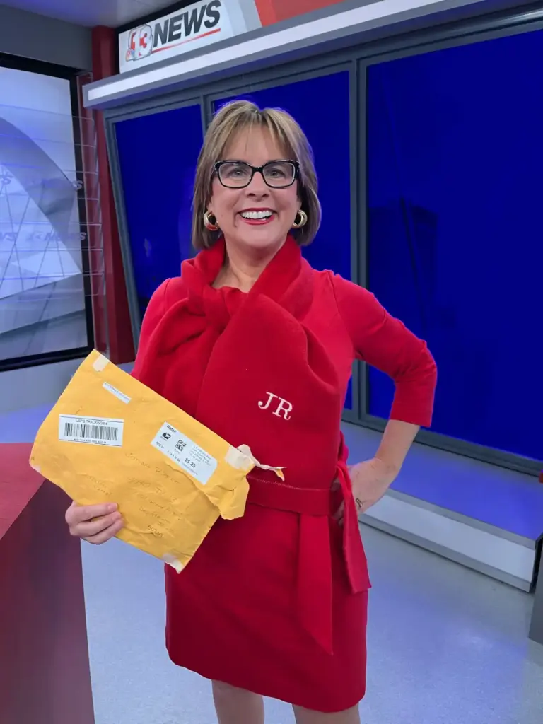 Meteorologist Jeriann Ritter in a red outfit holding a mailed package and wearing a personalized scarf in a TV studio.