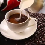 Coffee with milk being poured into a cup with roasted coffee beans, representing daily coffee consumption and its link to gut microbiome and cognitive effects