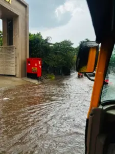 Flooded road in Pune with an auto rickshaw moving through rainwater during heavy unseasonal rainfall under overcast skies.