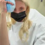 Taylor Momsen wearing a hospital gown and face mask, holding an ice pack to her head while seated in a medical room after a reported spider bite during her tour in Mexico City.