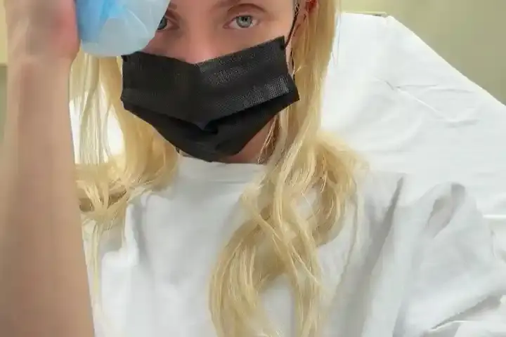 Taylor Momsen wearing a hospital gown and face mask, holding an ice pack to her head while seated in a medical room after a reported spider bite during her tour in Mexico City.