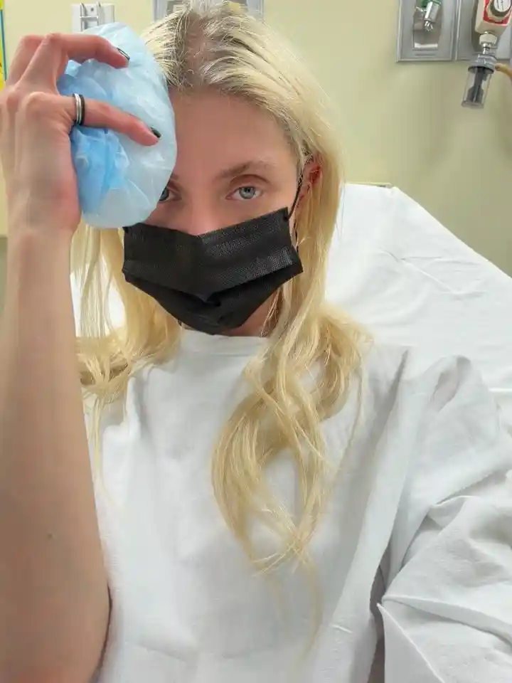 Taylor Momsen wearing a hospital gown and face mask, holding an ice pack to her head while seated in a medical room after a reported spider bite during her tour in Mexico City.