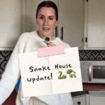 Callie holds a “Snake House Update!” sign inside her Montana farmhouse while discussing the ongoing garter snake issue linked to the home’s foundation.