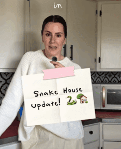 Callie holds a “Snake House Update!” sign inside her Montana farmhouse while discussing the ongoing garter snake issue linked to the home’s foundation.