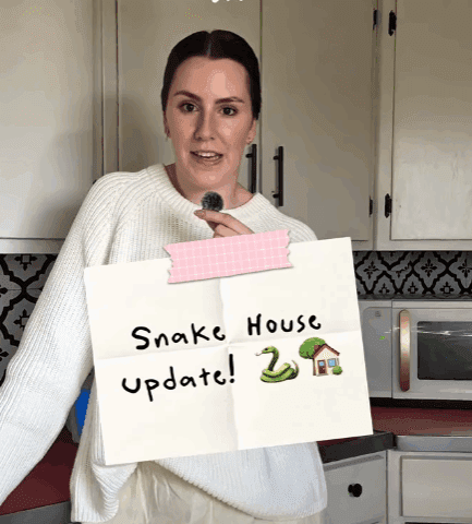 Callie holds a “Snake House Update!” sign inside her Montana farmhouse while discussing the ongoing garter snake issue linked to the home’s foundation.