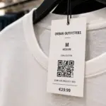 Clothing tag with a QR code and product details attached to a white cotton T-shirt, illustrating a Digital Product Passport concept for product transparency and traceability.