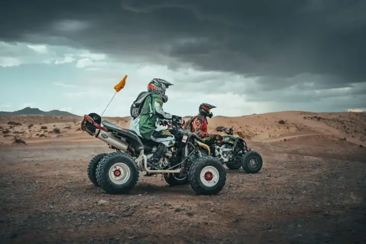 Two ATV riders wearing helmets and protective gear ride quad bikes across a desert landscape under dramatic clouds, representing an off-road ATV tour experience near Las Vegas.