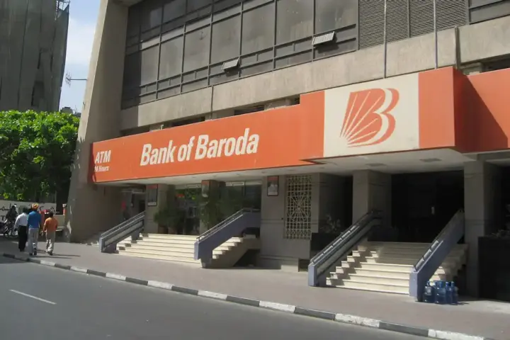 Exterior facade of a Bank of Baroda branch showing the bank's distinctive orange and white signage with the bank's logo, featuring multiple entrance steps and storefront entrance in an urban location.