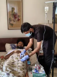 Healthcare worker wearing surgical mask and blue nitrile gloves administering intravenous fluid drip to a patient lying in a hospital bed during post-flooding disease outbreak, July 2026