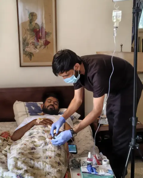 Healthcare worker wearing surgical mask and blue nitrile gloves administering intravenous fluid drip to a patient lying in a hospital bed during post-flooding disease outbreak, July 2026
