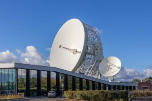 The Lovell 42-foot radio telescope at Jodrell Bank Observatory in Cheshire, UK, with its distinctive white dish antenna mounted on a steel lattice tower beside the SKA headquarters building, photographed against a blue sky with white clouds.