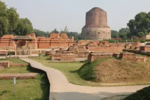 Sarnath archaeological site featuring the iconic red-brick Dhamek Stupa (circular cylindrical structure) and ancient Buddhist monument ruins set amid manicured lawns and surrounding trees, representing the sacred location where Buddha delivered his First Sermon.