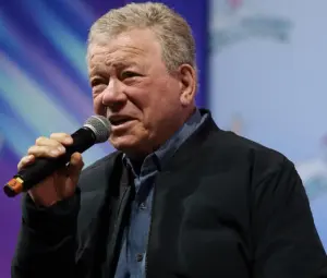 William Shatner, age 95, holding a microphone while speaking at a public event in December 2025, following his recovery from Stage 4 melanoma diagnosis.