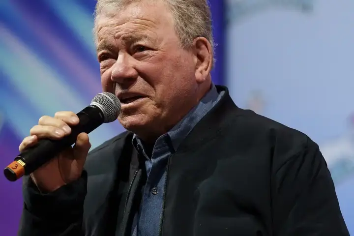 William Shatner, age 95, holding a microphone while speaking at a public event in December 2025, following his recovery from Stage 4 melanoma diagnosis.