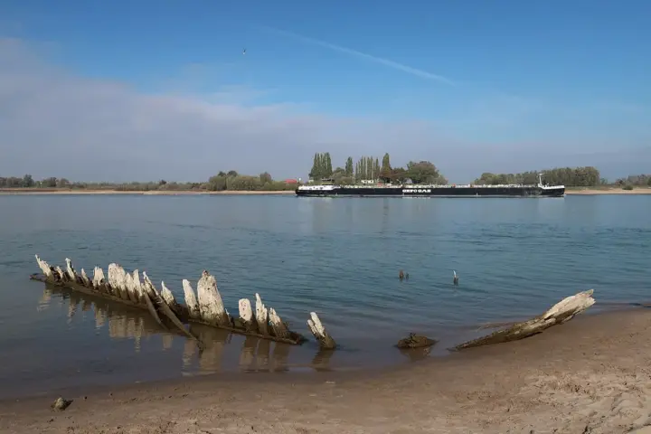 Exposed wooden ribs of a shipwreck on a sandy riverbank with low water behind it.