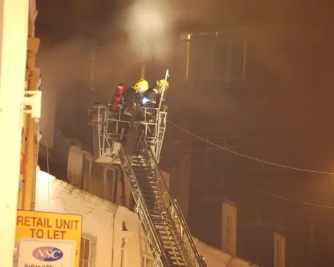 Firefighters in full gear operating on an extended aerial ladder platform against a smoke-filled building.