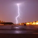 Dramatic thunderstorm with bright lightning striking over an airport tarmac at dusk or dawn. Several aircraft are parked near a brightly lit terminal building; wet pavement reflects the storm light. The image captures the severe weather conditions that disrupted flight schedules across Northeast US airports.