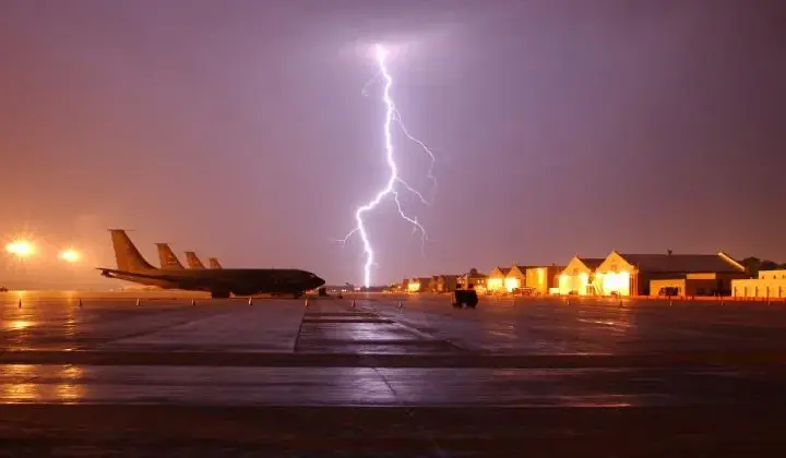 Dramatic thunderstorm with bright lightning striking over an airport tarmac at dusk or dawn. Several aircraft are parked near a brightly lit terminal building; wet pavement reflects the storm light. The image captures the severe weather conditions that disrupted flight schedules across Northeast US airports.