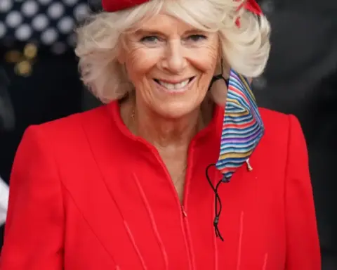 Queen Camilla of the United Kingdom smiling in a red coat and matching hat at a public engagement.