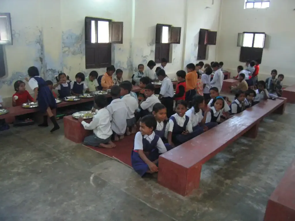 Rural Indian schoolchildren seated on benches eating a midday meal of rice and curry in a school hall.