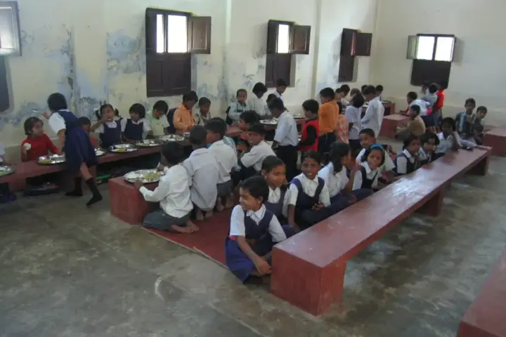 Rural Indian schoolchildren seated on benches eating a midday meal of rice and curry in a school hall.