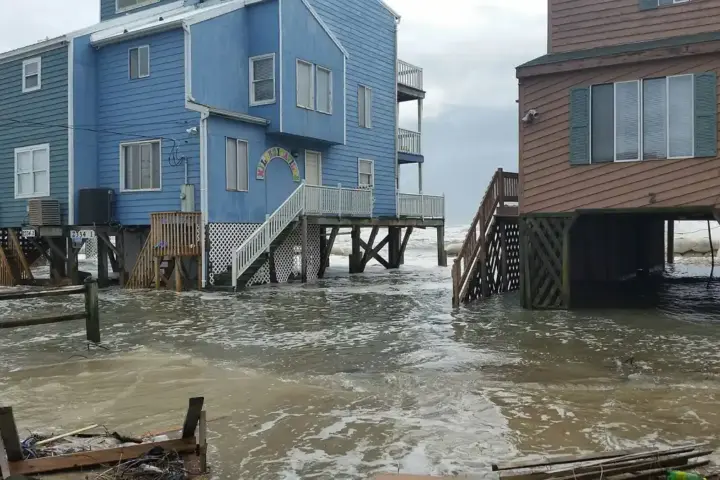 Floodwater covering the ground beneath stilt houses in a coastal North Carolina neighbourhood, with debris floating in murky brown water under an overcast sky