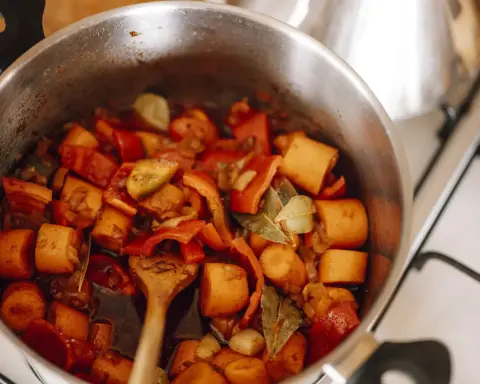 Chopped carrots, red peppers, onions and two bay leaves simmering in dark broth in a stainless steel pot on a domestic stovetop, with a wooden spoon at the edge
