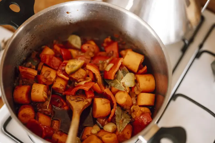 Chopped carrots, red peppers, onions and two bay leaves simmering in dark broth in a stainless steel pot on a domestic stovetop, with a wooden spoon at the edge