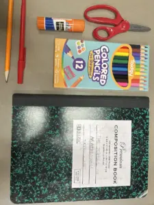 School supplies arranged on a grey surface including a yellow pencil, red pen, glue stick, scissors, a boxed set of twelve coloured pencils and a composition notebook