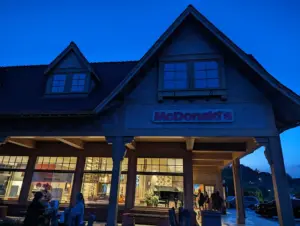 Exterior of a McDonald's restaurant at twilight with lodge-style timber pillars and a shingled roof, golden arches sign lit above large windows and customers seated outside