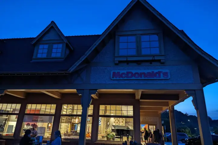 Exterior of a McDonald's restaurant at twilight with lodge-style timber pillars and a shingled roof, golden arches sign lit above large windows and customers seated outside
