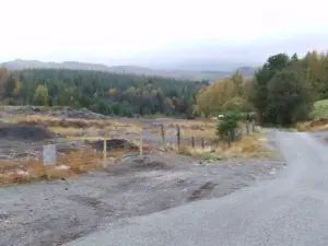Fenced-off area of stripped dark earth beside a rural road, with forested hillside behind under an overcast sky