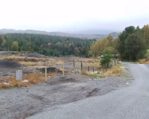 Fenced-off area of stripped dark earth beside a rural road, with forested hillside behind under an overcast sky