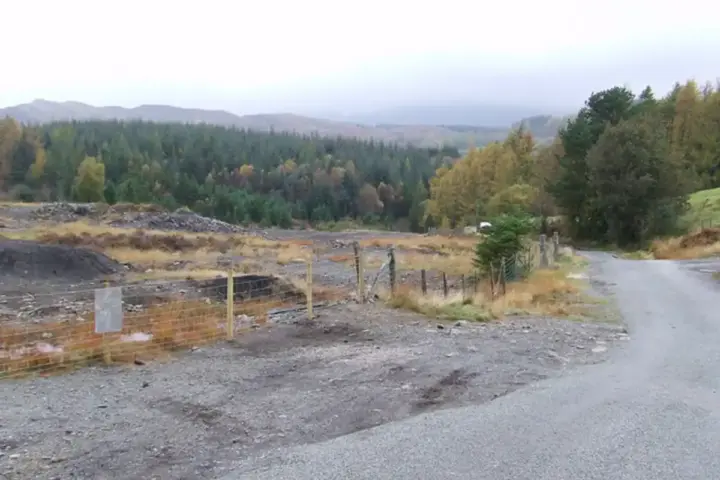 Fenced-off area of stripped dark earth beside a rural road, with forested hillside behind under an overcast sky