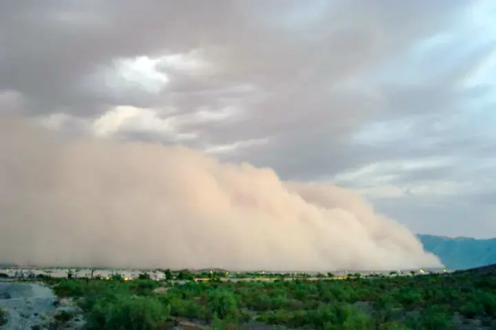 A wall of dust advances toward a lit desert highway corridor with mountains behind.