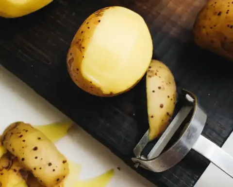 Close-up of roasted potatoes with golden-brown, speckled skin, showing a peeled slice revealing yellow interior, with a metal peeler nearby