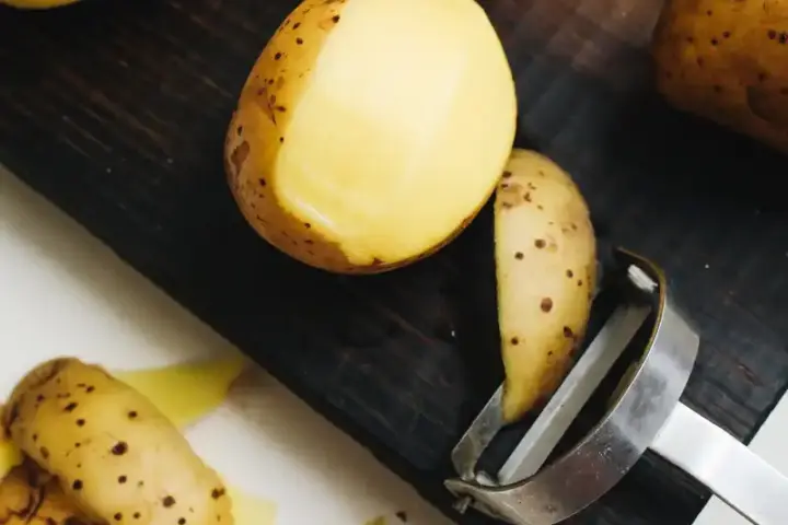 Close-up of roasted potatoes with golden-brown, speckled skin, showing a peeled slice revealing yellow interior, with a metal peeler nearby